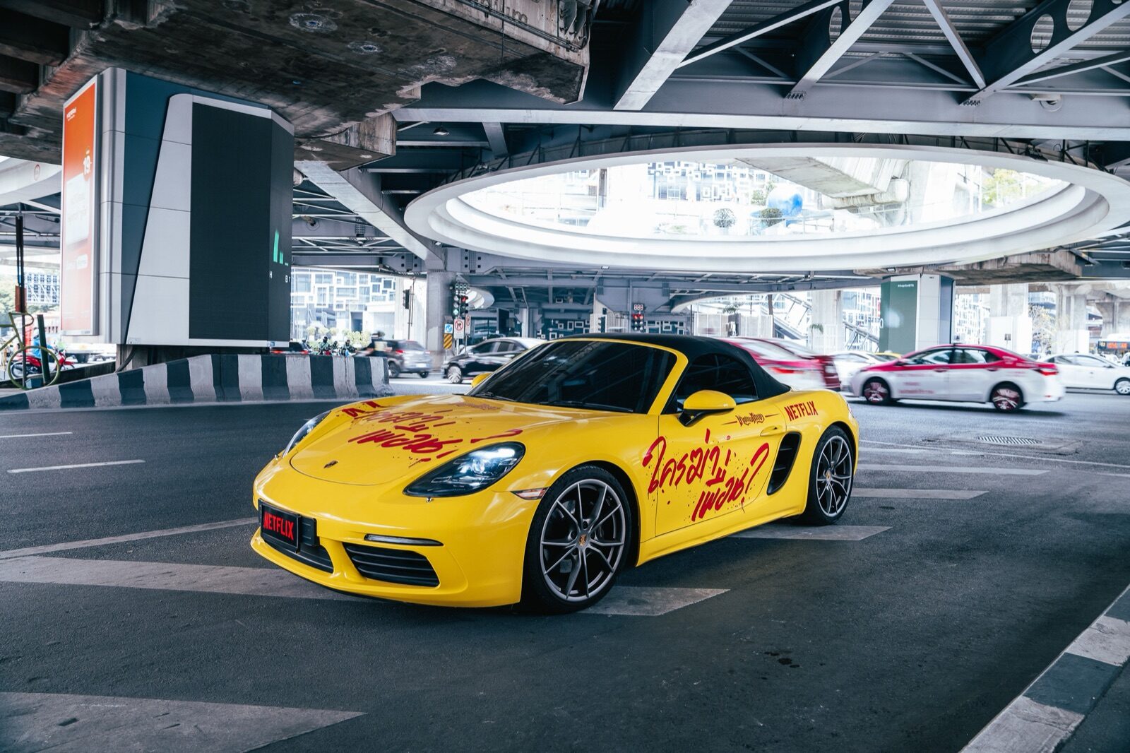 A yellow Netflix-wrapped Porsche parked under a dramatic Bangkok expressway interchange